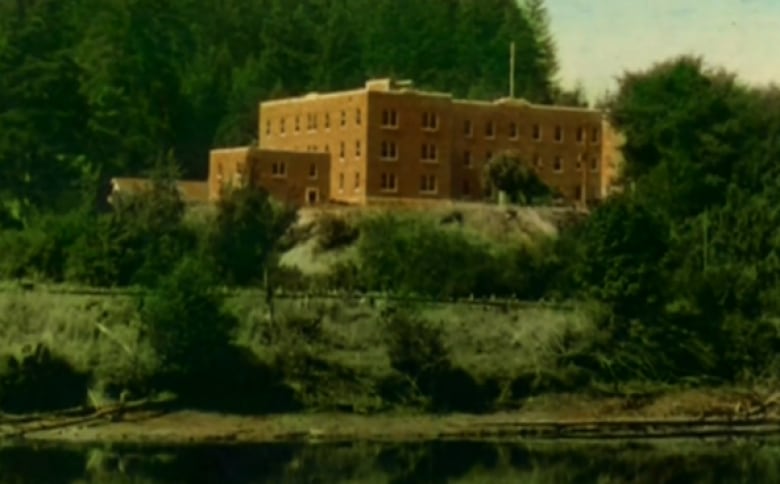 An old colour photograph of a brick building on a cliff surrounded by trees over water.