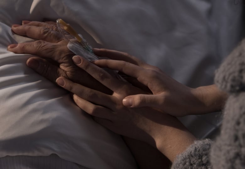 The hands of an adult hold the hand of an older person, who is hooked up to intravenous and lying in a bed.