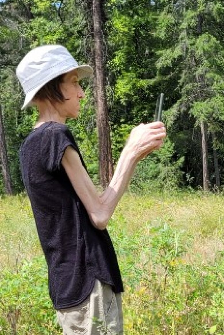 A woman wearing a hat looks at her smartphone while standing in an overgrown field with trees behind her.