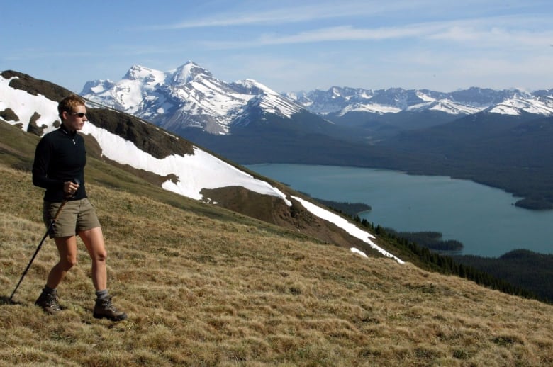 A person hiking in shorts and a long-sleeved top and carrying a staff walks along a grassy mountain. Snow-capped mountains and a lake are in the background.