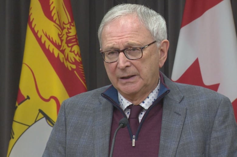An older man with a furrowed brow standing in front of a New Brunswick flag and a Canada flag.