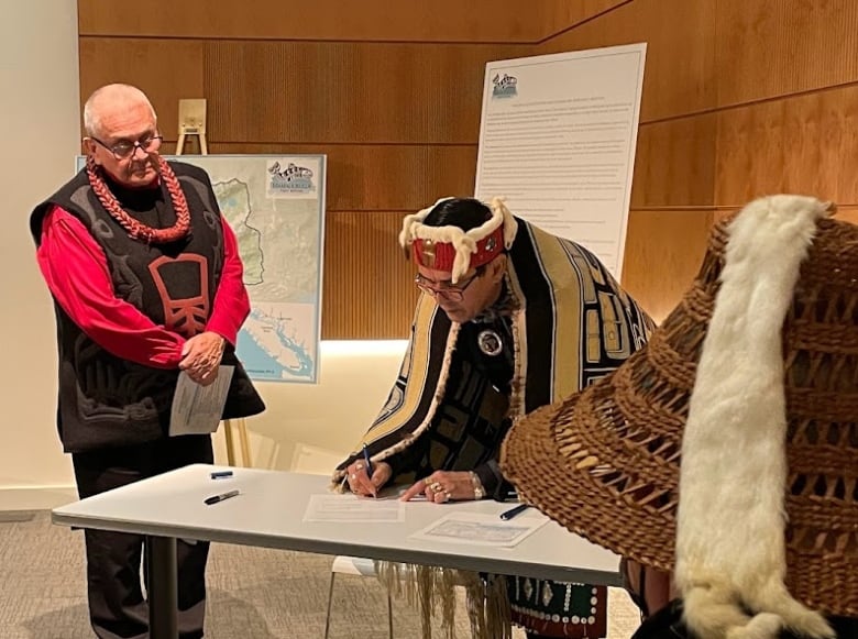 A man in traditional Indigenous regalia signs an agreement while others watch in a meeting room.