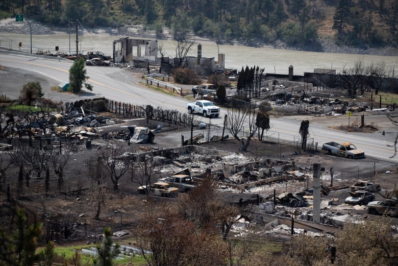 A white police truck drives down a road past shells of burned-out vehicles and structures destroyed by a wildfire. 