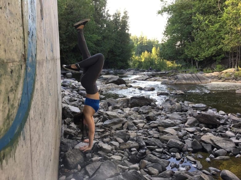 A woman does a handstand on the rocks near a creek and forest. 