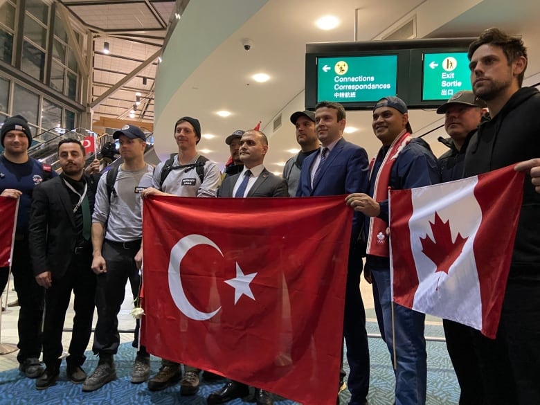 A man in a suit holds a Turkish flag, surrounded by a crowd of men mostly wearing clothes to work in. Another man holds a Canadian flag.
