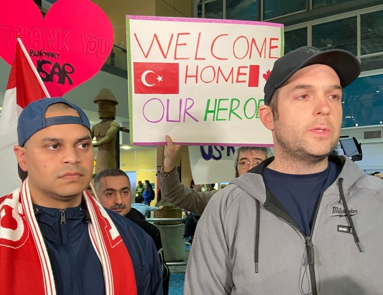 Two men, one wearing a CANADA scarf, speak to a reporter in the airport while a sign reading "Welcome Home Our Heroes" is held in the background
