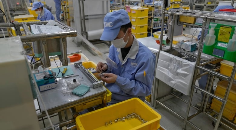 A man wearing a blue uniform and a face mask assembles machinery parts in a factory.