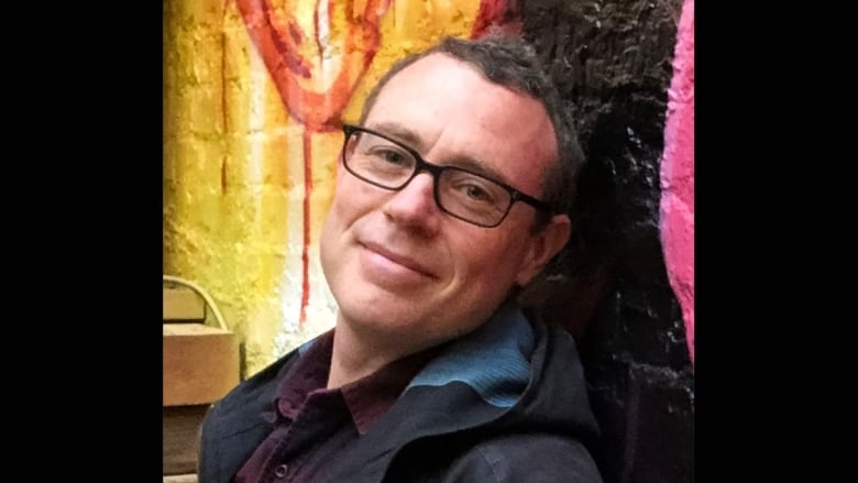A smiling white man wearing dark-rimmed glasses leans against a painted brick wall. He is wearing a dark purple collar shirt and a dark hooded zip-up jacket.