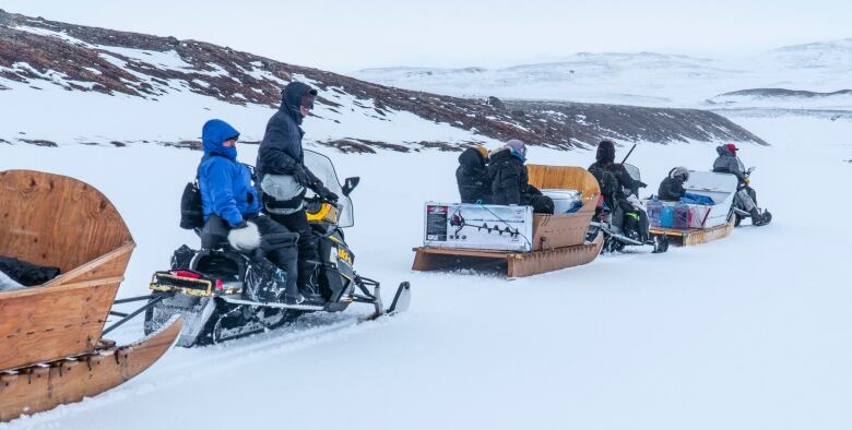 A line of people on skidoos pull wooden sleds loaded with people and supplies for ice fishing.