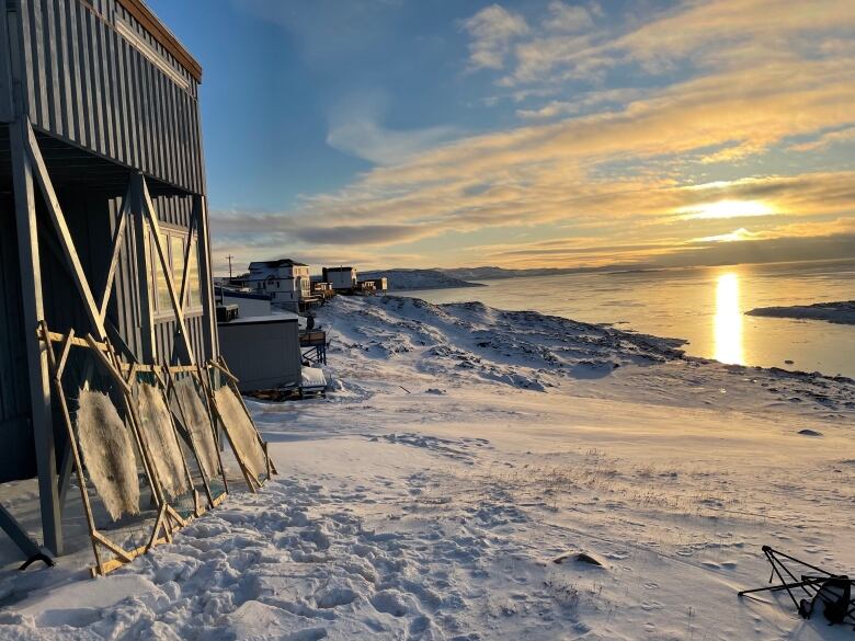 The Arctic sun glows across the water as four sealskins, stretched in frames to dry, lean against a building.