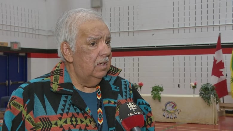 A man in blue ceremonial wear is speaking into a CBC microphone.