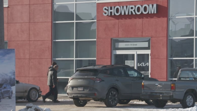 Two men — a KIA salesman and Go Public's secret shopper — enter KIA West Edmonton car dealership. The exterior of the dealership is red. Both men are wearing coats. 