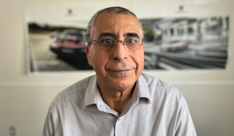A brown man is wearing glasses and is sitting in front of two posters of luxury vehicles. The posters are blurry while he is in focus. 