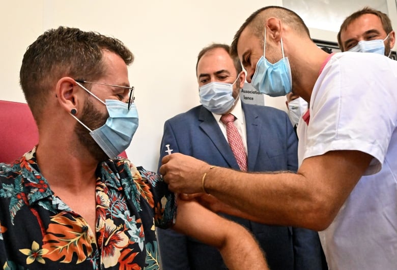 Person wearing tropical short-sleeved shirt and medical face mask sits as a health professional wearing a mask holds vaccine needle above his arm, while others watch.