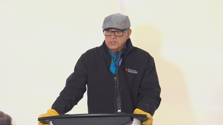 A man wearing a grey cap, a black jacket and yellow gloves speaks at a podium.