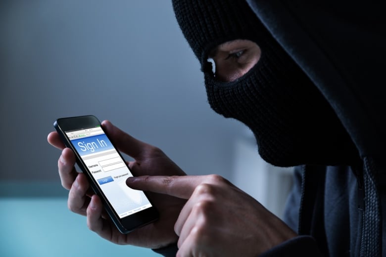 A stock image of a hacker in a full face covering signing into an app on a phone.
