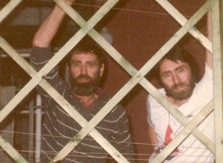 Two men stand behind a cross-hatched fence.