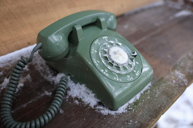 An olive green rotary phone on top of a wooden table surrounded by a dusting of snow.