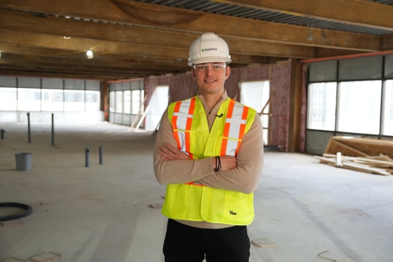 A man in a yellow reflective safety vest and white hard hat stands inside a big open concept office space that's being revovated. 