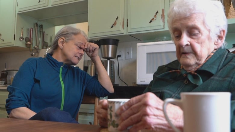 A 60-year-old woman on the left with her hand on her forehead sits at a table next to a 92-year-old woman holding a mug of tea.