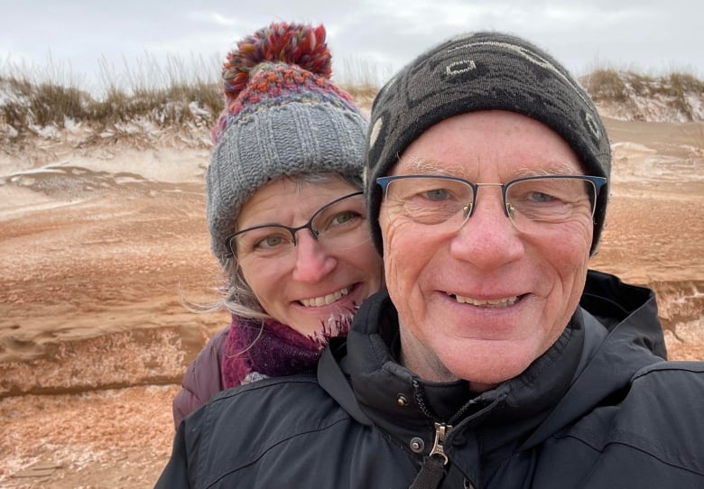 A man and a woman dressed in winter coats and toques smile for the camera in front of a backdrop of sand dunes.