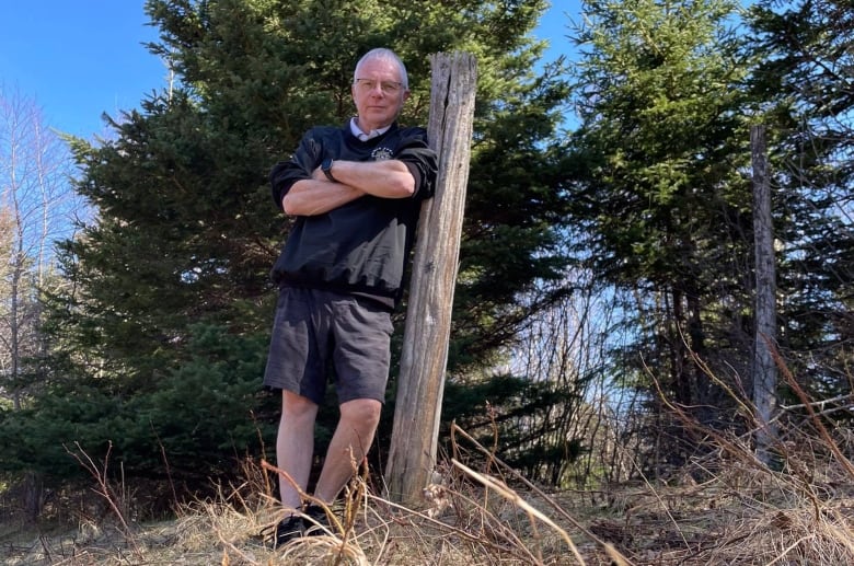 A man in shorts and a sweater leans against a wooden post, surrounded by trees.