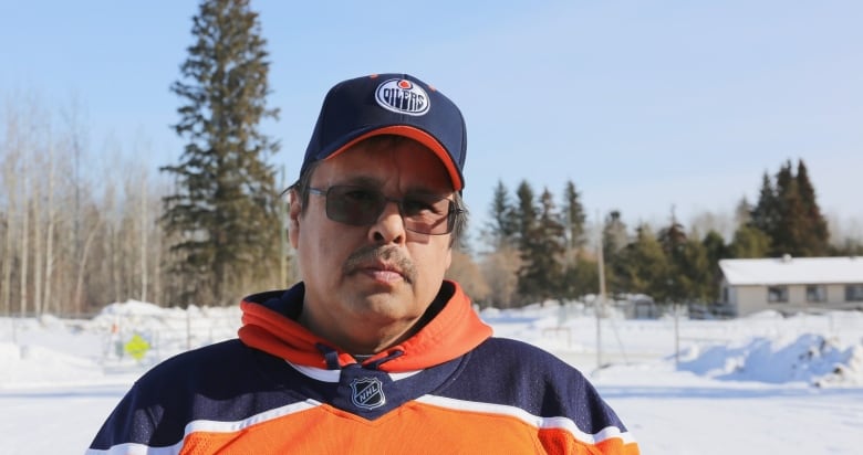 A man in dark glasses stands outdoors in winter, wearing an Edmonton Oilers jersey and cap.