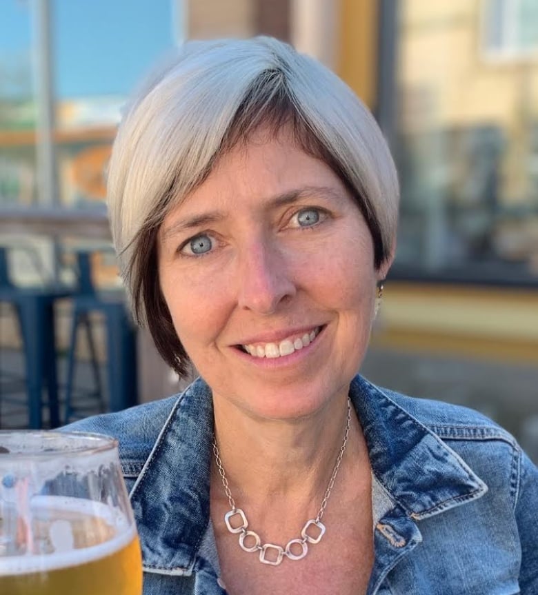 a woman wearing a jean jacket smiles and holds a pint of beer. she has short white hair 