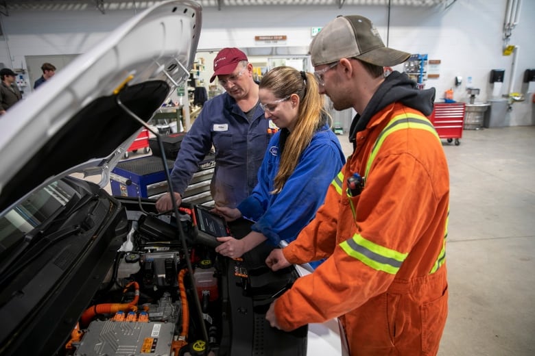 A coveralls-clad instructor and students, one female and one male, lean over an opened Chevy Bolt hood to peer at a tablet connected to the electric vehicle's components.