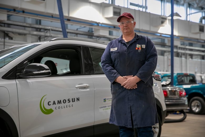 A man in a burgundy cap and navy coveralls stands in a bright and spacious college auto body shop, next to a white vehicle with a logo for Camosun College on its door. 