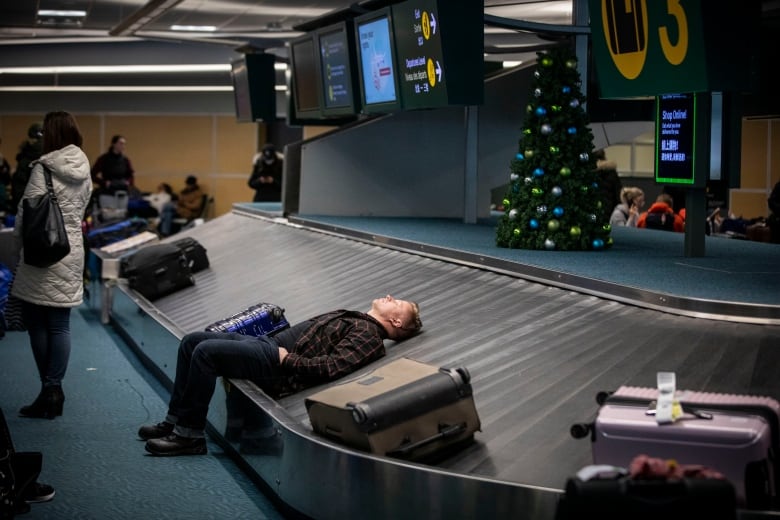 A man with a suitcase beside his head lies down and closes his eyes on a baggage carousel with a Christmas tree in the background and passengers milling around.
