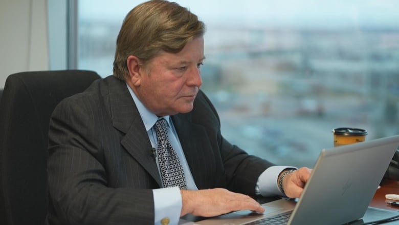 A man with brown hair, wearing a suit and tie, sits at a desk in front of an office window, looking at a laptop screen.