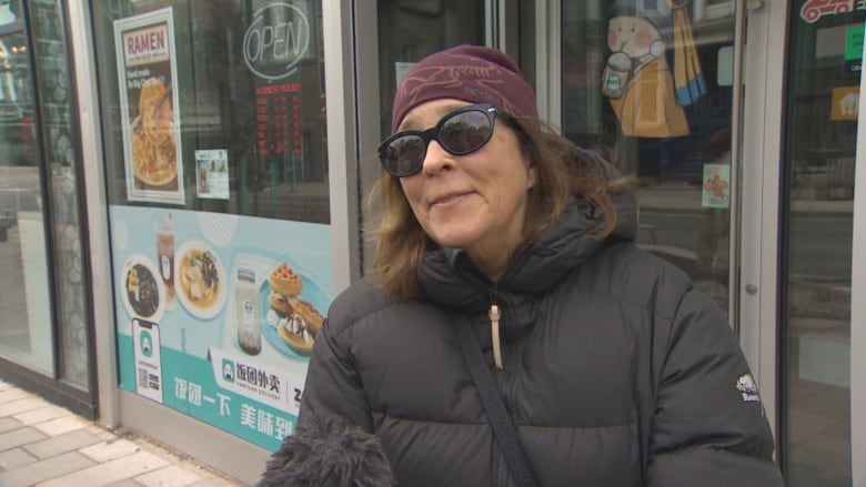 A woman in a purple toque, sunglasses and a black parka stands on a sidewalk in front of a ramen restaurant.