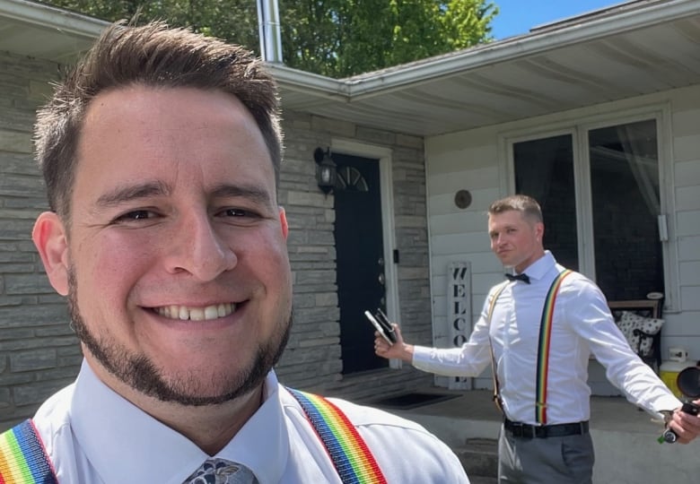 Two men wear white shirts and rainbow suspenders in front of a house. 