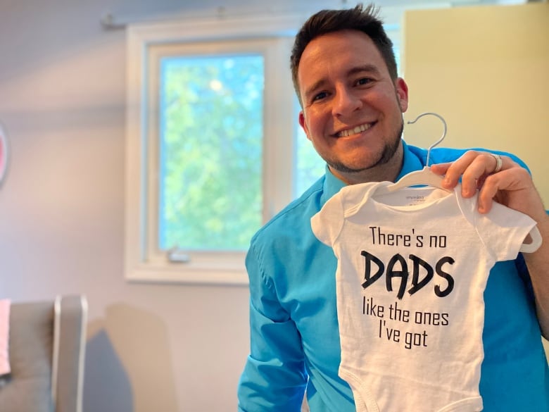 A man holds a onesie meant for a newborn. The printed letters on it read "There's no dads like the ones I've got."