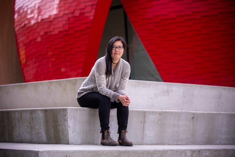 A women in a grey sweater in black pants and glasses sits on concrete stairs in front of a red building.