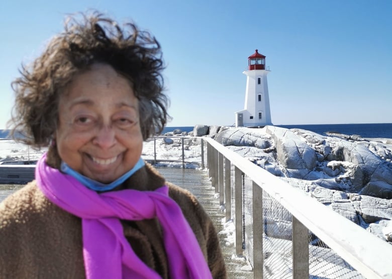 A woman with short, dark and wavy hair stands smiling outside, with a lighthouse in the background to the right of the photo. She is wearing a light brown coat and pink scarf tied loosely around her neck.