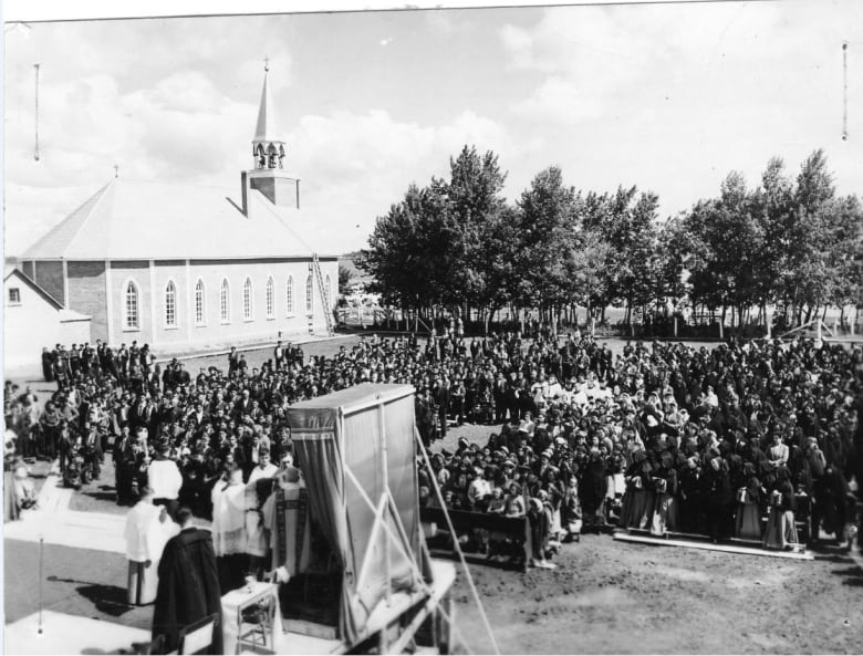A black and white photo shows a large crowd on the grounds of the old boarding school in Île-à-la-Crosse.