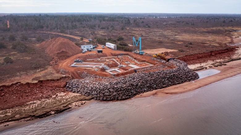 Drone view of a huge rock breakwater being built around a new home on a red-sand shoreline.