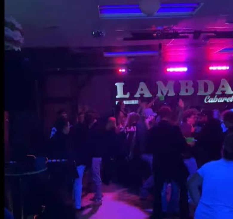 A video still shows a number of people dancing in a dark club in front of a sign that reads "Lambda."