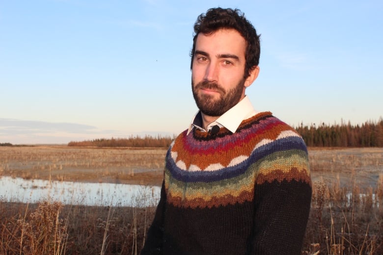 A man with brown hair and a beard stands is wearing a sweater with a coloured pattern near the neck. He stands in front of mine tailings, the waste left over from a gold mine in Val D'Or. There are shallow pools of water and some browning plants growing from the waste.