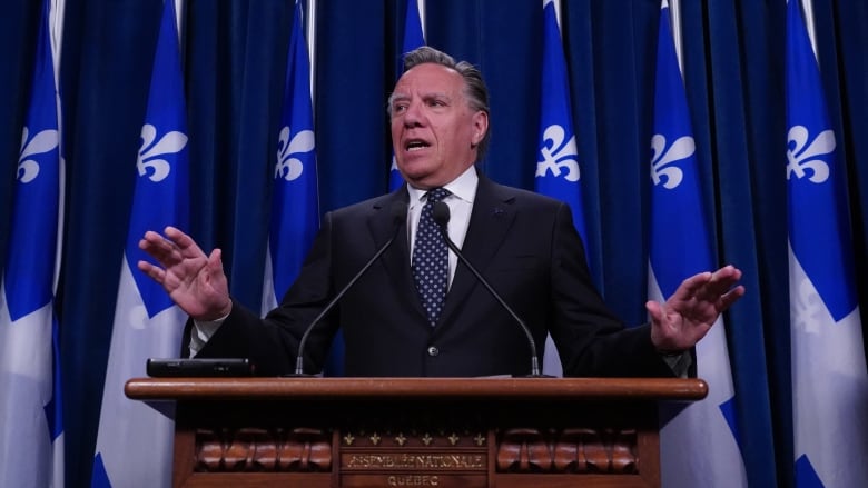 A man speaking at a podium in front of Quebec flags. 