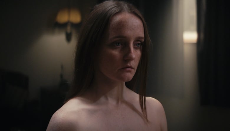 A still from a film shows a sad-looking woman with bare shoulders standing in a dimly lit room of a home.
