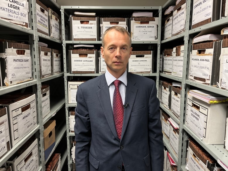 A man in a suit standing in a room with filing boxes.