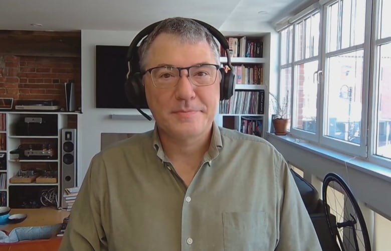 A man in a beige shirt, wearing a headset, in a living room, looks straight at the camera.