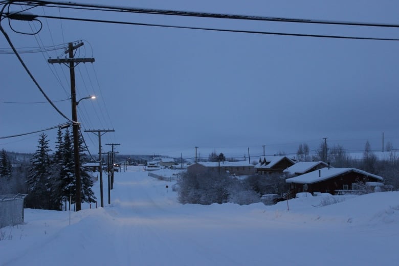 A photo taken from a road, with power lines on the left and looking down on snow-covered homes to the right and in the distance. It's dawn and there's isn't a lot of light - and street lights are on.