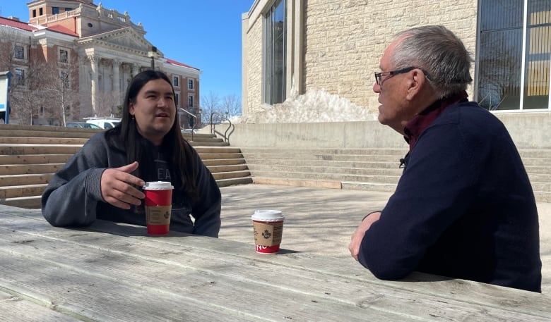 A student sits at a bench outside of his University with a coffee and is having a conversation in Cree
