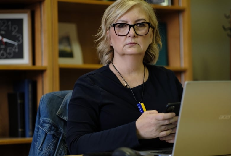 A woman wearing glasses and a dark sweater, and with a serious expression on her face, sits at a desk working on a computer.