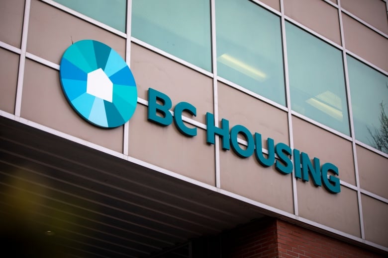 A blue circular symbol, with a house within it, next to a sign that reads 'BC Housing' is affixed on a building.