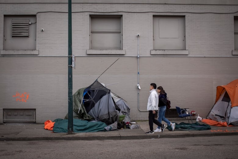 Two people walk past tents on a city sidewalk.
